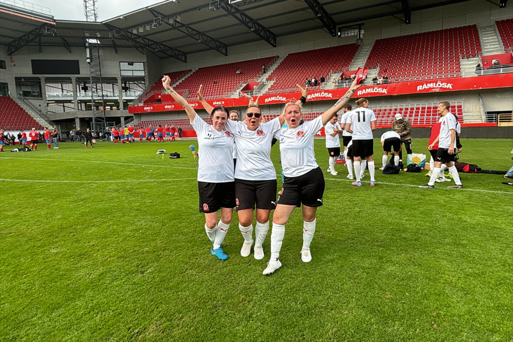 Tre kvinnliga fotbollsspelare på fotbollsplanen i en fotbollsarena, som håller upp sina armar och tjoar.